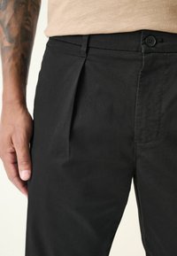 Pantalon plissé noir avec une texture lisse, des passants de ceinture, une fermeture à bouton et une poche latérale, placé juste au-dessus d'une main tatouée.