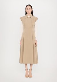 Rochie bej fără mâneci, cu guler, având pliseuri verticale și o fustă evazată. Purtați-o peste un top cu mâneci lungi, cu o tekstură fină.