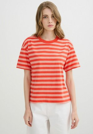 Joven mujer con cabello rubio ondulado, que lleva una camiseta de manga corta a rayas rojas y blancas y pantalones blancos, de pie frente a un fondo blanco.