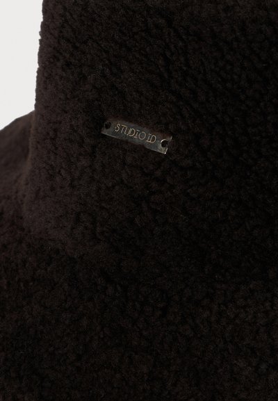 Donkerbruin textuur fleece stof met een klein metalen label met de tekst "STUDIO ID" dat aan het oppervlak is bevestigd.