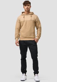 Sudadera beige a rayas con un bolsillo frontal, cordones y puños acanalados, combinada con pantalones negros ajustados y zapatillas blancas.