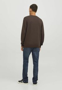 Camisa marrón de manga larga con puños y dobladillo acanalados, combinada con jeans de mezclilla azules. El modelo lleva zapatillas negras con suela blanca.