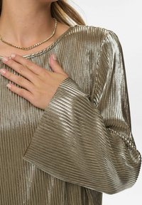 Metalicky strieborný top s vertikálnymi plisovaním, širokými rukávmi a zaobleným výstrihom. Ruka modelu spočíva na materiáli. Viditeľný textúrovaný lesk.