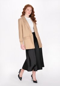 Beige blazeri, jossa on pitkät hihat ja kaksi etutaskua, yhdistetty mustaan satiiniseen midi-hameeseen ja mustiin korkeakorkoisiin kenkiin.