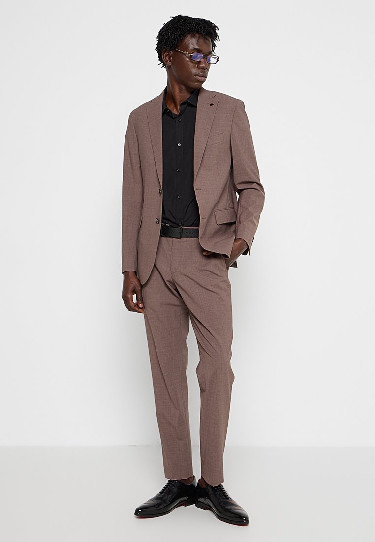 Abito marrone con tessuto strutturato, blazer a un bottone, camicia nera sotto, abbinato a scarpe nere. Taglio slim-fit, linee pulite.