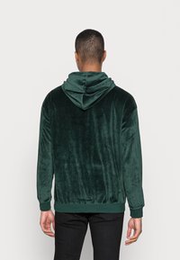 Grön sammets-huvtröja med dubbel-lager huva, ribbade ärmslut och nederkant. Slät textur, avslappnad passform och minimalistisk design.