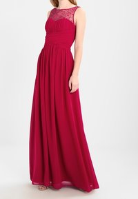 Lång formell klänning i djup röd med en figursydd liv, plisserad kjol och en genomskinlig, pärlbeströdd halsringning. Mjuk, flödande tyg.