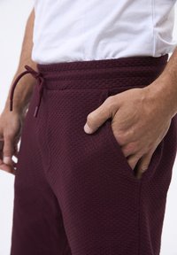 Burgunderfarvede joggers med struktur, elastisk liv og justerbar snøre, med sidelommer, vist i taljehøjde.