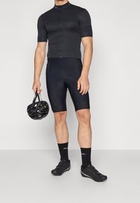 Maillot et cuissard de cyclisme noirs en tissu extensible, associés à des chaussures de cyclisme noires et à un casque, le tout dans un design moderne et épuré.