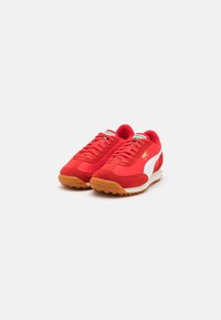 Red athletic sneakers made of suede and mesh, featuring a white logo, gum rubber outsole, and flat laces. Minimalist design with reinforced toe.
