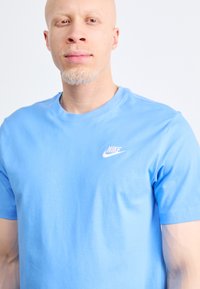 Nebesno modra majica s kratkimi rokavi iz bombaža, ki ima belo Nike logo na levem prsnem delu. Gladka tekstura in krojen ovratnik.