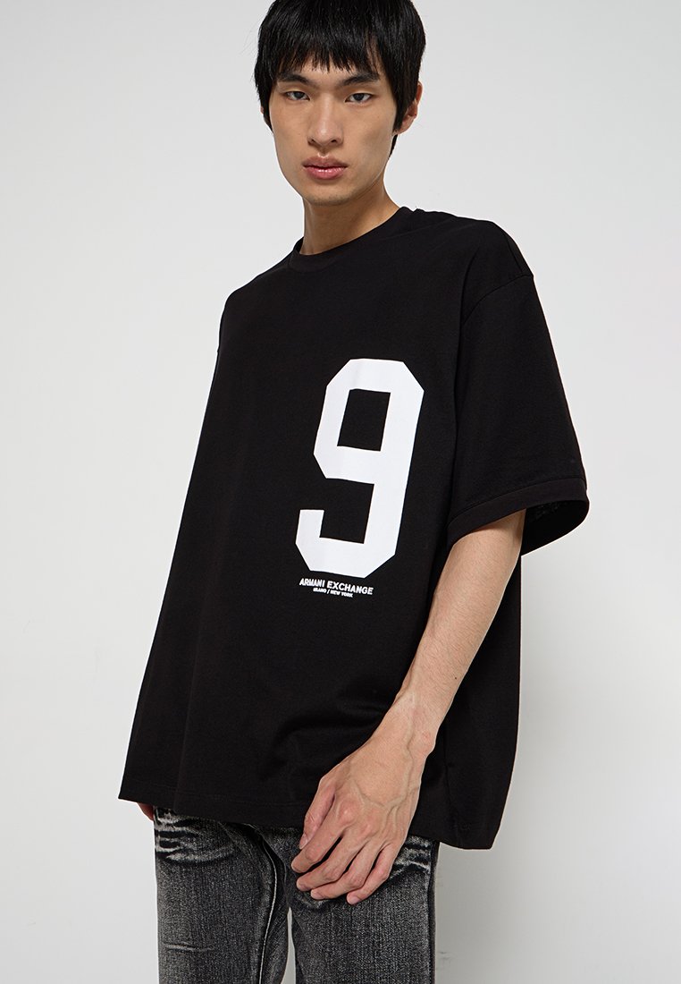 Black oversized T-shirt featuring a large white number "9" on the front and "Armani Exchange" logo at the bottom. Cotton blend fabric.