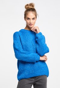Oversized blue knitted sweater with ribbed texture and subtle metallic threads. Wide sleeves and crew neckline. Paired with gray pants.