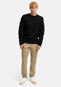 Schwarzer Strickpullover mit einem strukturierten geometrischen Muster, Rundhalsausschnitt und gerippten Bündchen, kombiniert mit beigen Chinos und weißen Sneakers.