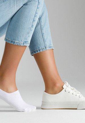 Sneaker in tela bianca con suola in gomma chiara accanto a un semplice calzino alla caviglia bianco, su sfondo neutro, che mostra uno stile casual.