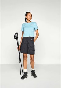 Lys blå avkortet t-skjorte, svarte cargo-shorts med flere lommer, svarte tursko og stav for fotturer mot en nøytral bakgrunn.