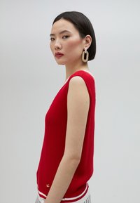 Röd ribbad ärmlös topp med vit kant och en guldlogotypdetalj. Modellen bär stora cirkulära örhängen mot en neutral bakgrund.