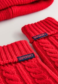 Røde strikkede fingeløse handsker med ribbet manchet, med et kabelstrikket mønster og et blåt "SUPERDRY" mærke på siden.