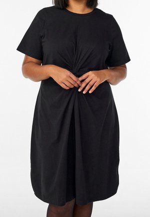 Donna che indossa un vestito nero a maniche corte con dettaglio nodo frontale, in piedi su uno sfondo bianco.