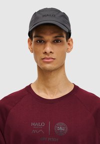 Schwarze Kappe mit dem "HALO"-Logo, ausgestattet mit einem gebogenen Schirm und einem leichten, strukturierten Stoff. Getragen mit einem weinroten Sweatshirt, das grafische Prints zeigt.