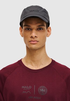 Zwarte pet met "HALO" logo, voorzien van een gebogen rand en een lichte, gestructureerde stof. Draagt samen met een bordeauxrode sweatshirt met grafische prints.