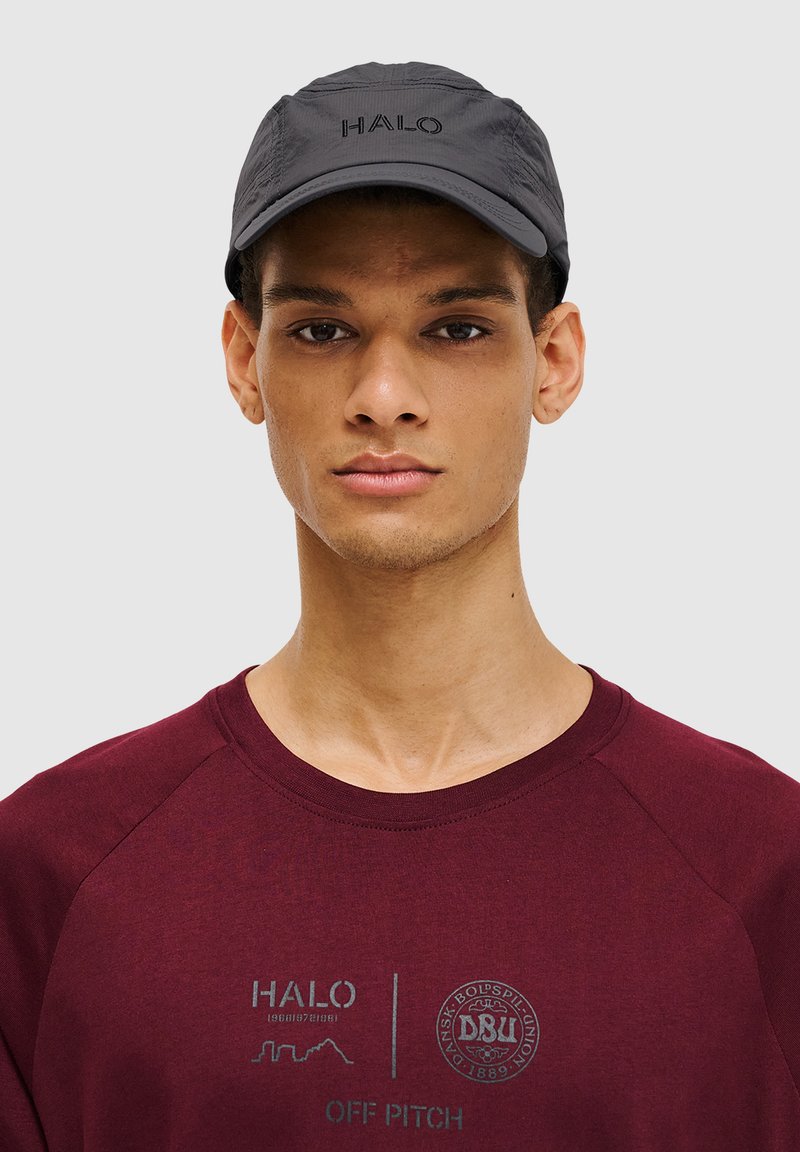 Schwarze Kappe mit dem "HALO"-Logo, ausgestattet mit einem gebogenen Schirm und einem leichten, strukturierten Stoff. Getragen mit einem weinroten Sweatshirt, das grafische Prints zeigt.