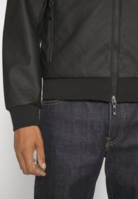 Giacca bomber nera trapuntata con zip frontale, polsini e orlo a costine. Indossata sopra jeans in denim scuro con dettagli di cucitura gialla.