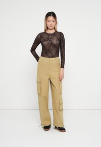 Top lungo a maniche trasparenti marrone con motivo floreale; pantaloni cargo beige con tasche laterali; la modella indossa scarpe con piattaforma nere su uno sfondo bianco.