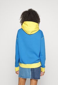 Sudadera con capucha azul y amarilla, con un bolsillo canguro frontal, puños y dobladillo acanalados, combinada con una falda mini de mezclilla.