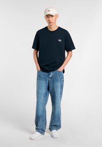 Navy crew-neck t-paita, jossa on pieni logo, yhdistettynä vaaleansinisiin rentoihin farkkuihin. Valkoiset lenkkarit ja beige lippalakki täydentävät asustusta.