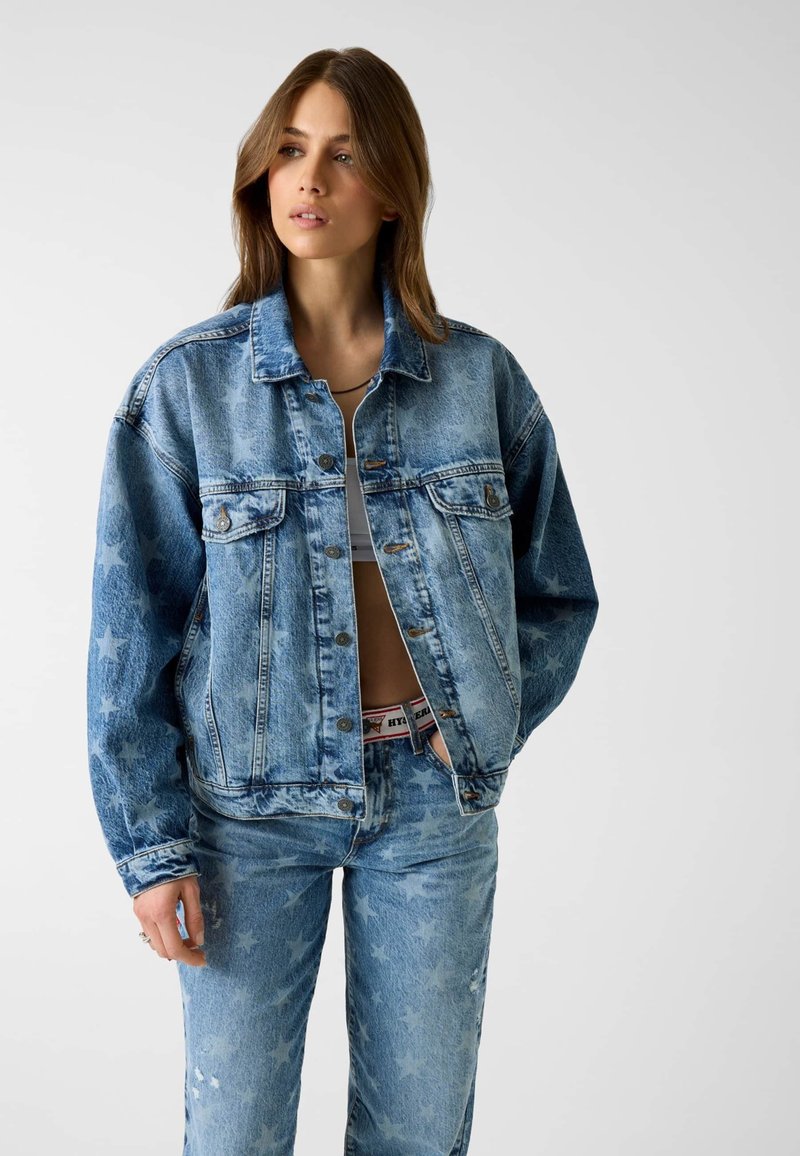 Femme portant une veste en denim bleu assortie et un jean avec des motifs d'étoiles, un crop top blanc et une ceinture avec une taille visible.