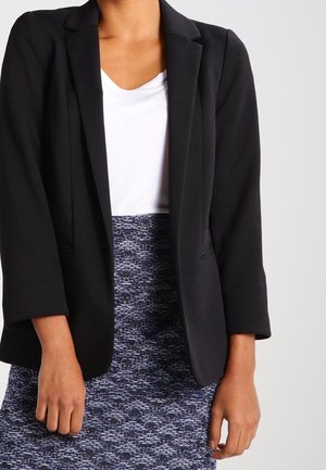 Person som bär en svart blazer, vit topp och blåmönstrad kjol, står med armarna avslappnat vid sidorna mot en neutral bakgrund.