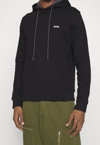 Sweat à capuche en coton noir avec une poche frontale, des cordons ajustables et un petit logo blanc "BOSS". Associé à un pantalon cargo vert olive avec des fermetures éclair.