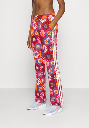 Personne portant un pantalon de survêtement Adidas à motif floral coloré avec des rayures blanches et des baskets Adidas bleu clair, debout contre un mur blanc.