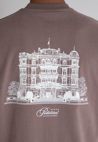 Brown t-shirt featuring a white outline illustration of an elegant building with trees and the text "Prohibited Grand Resort" below.