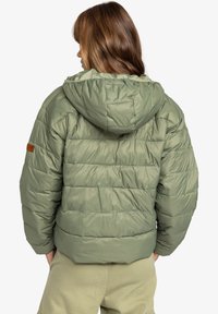 Olivengrønn pufferjakke med hætte, quiltet design, elastiske manchetter og et mærke på venstre ærme. Glat, let skinnende tekstur.