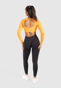 Langärmliges orangenes Cropped Top mit einem runden Ausschnitt auf dem Rücken, kombiniert mit hochtaillierten schwarzen Leggings und weißen Sneakers.