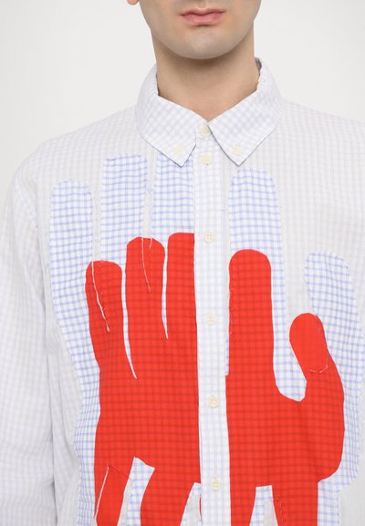Camisa de cuadro con botones que presenta grandes huellas de manos abstractas rojas sobre un fondo blanco, con costuras visibles y textura de la tela.