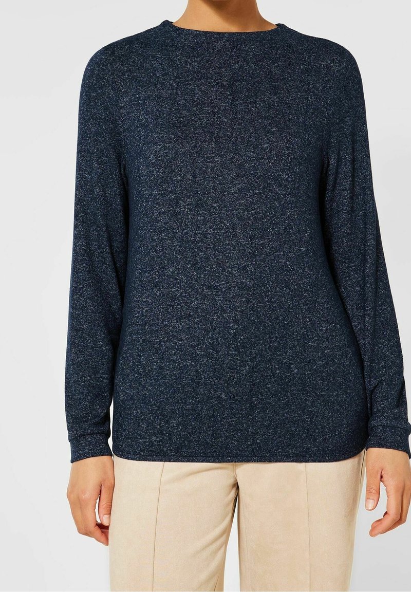Personne portant un haut en tricot à manches longues bleu foncé et un pantalon beige, debout devant un fond clair uni.