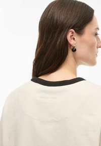 Ženska z dolgimi rjavimi lasmi, ki nosi srebrne uhančke obroče s črnimi kroglicami ter bež srajco s črnim ovratnikom in napisom "ROUGE EDIT".