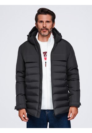 Homme portant une doudoune noire avec capuche par-dessus un sweat à capuche blanc zippé et un jean bleu sur un fond clair uni.