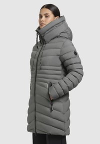 Donna che indossa un lungo piumino grigio con colletto alto e cappuccio, con le mani nelle tasche con cerniera, su uno sfondo uniforme.