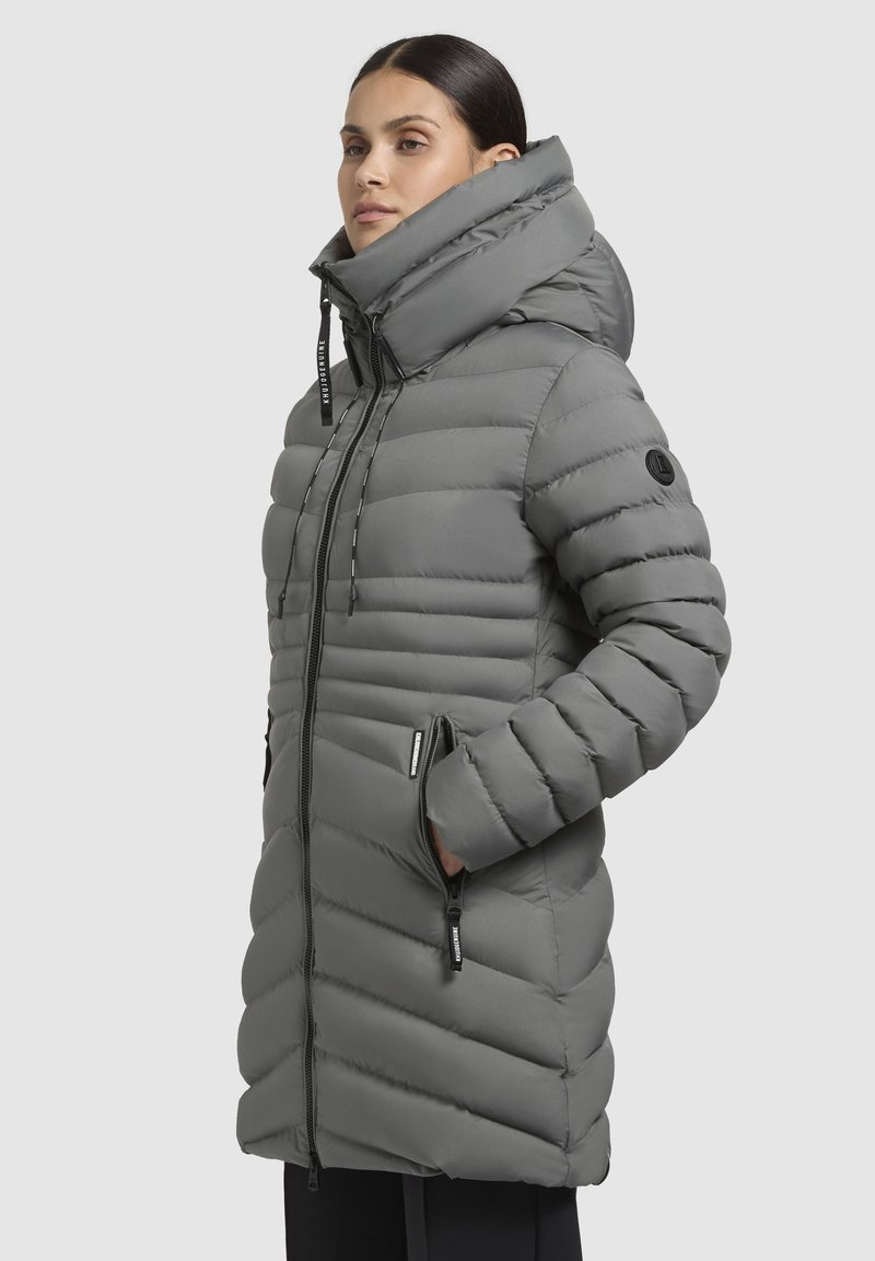Donna che indossa un lungo piumino grigio con colletto alto e cappuccio, con le mani nelle tasche con cerniera, su uno sfondo uniforme.