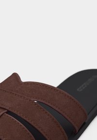 Gros plan sur une bride de sandale en daim marron sur une semelle noire avec des coutures visibles et un logo de marque flou.