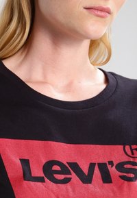 Černé tričko s krátkým rukávem, na kterém je velké červené logo Levi's v tučném černém písmu. Měkký bavlněný materiál s kulatým výstřihem.