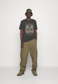 Camiseta gráfica gris con diseño de serpiente, combinada con pantalones cargo oliva y un sombrero tipo bucket bicolor. Zapatos marrones y corte relajado.