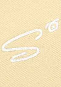 Hellgelber Stoff mit einem strukturierten Strickmuster, das weiße, gestickte Kursive in einem lockeren, fließenden Stil aufweist.