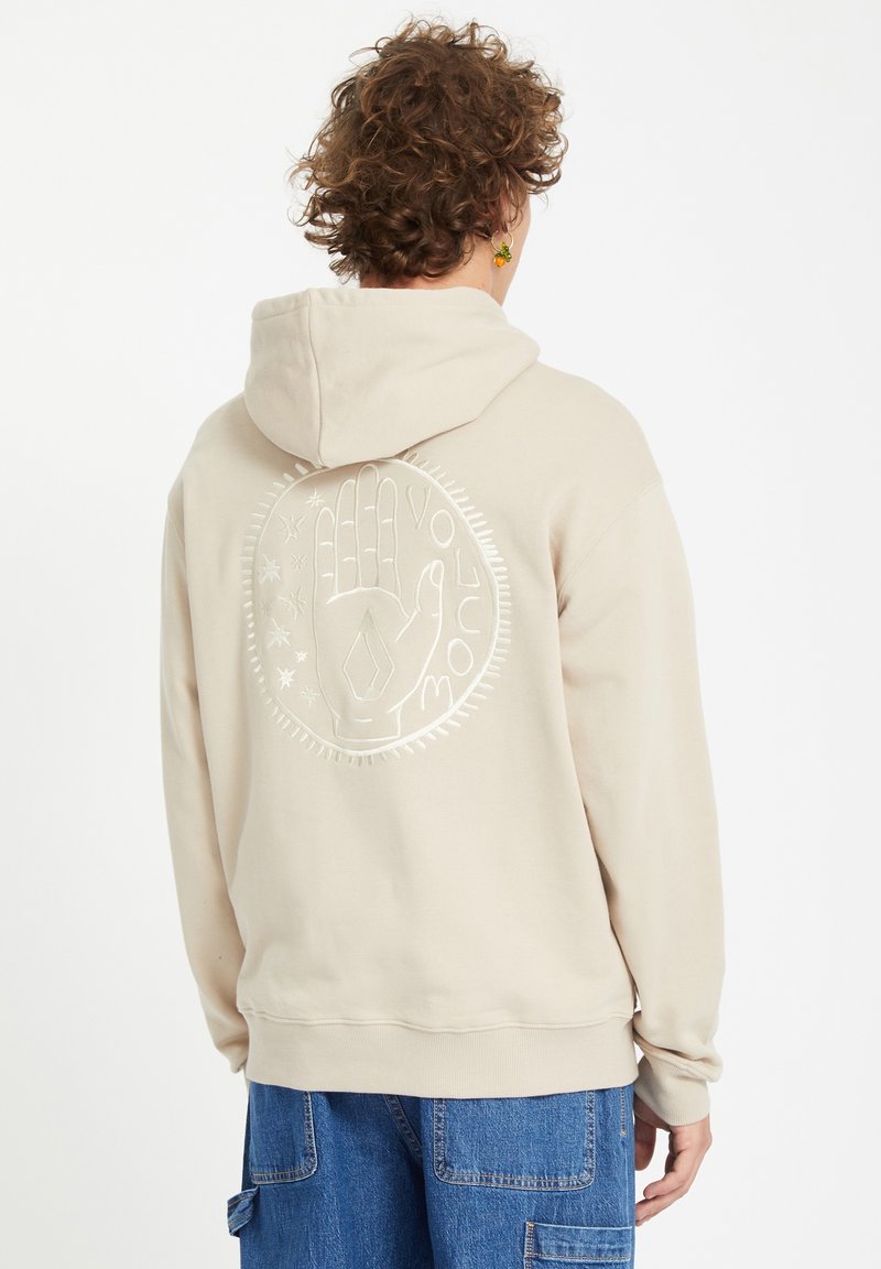Personne aux cheveux bouclés portant un sweat à capuche beige avec un design en relief représentant une main circulaire et des étoiles, accompagné du texte « Moujoco », associé à un jean bleu.