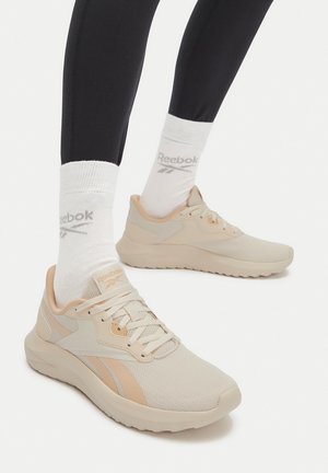 Pies con zapatillas Reebok beige y calcetines blancos Reebok combinados con leggings negros sobre un fondo claro y sencillo.