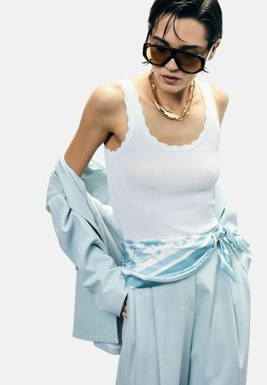 Donna con grandi occhiali da sole, collana con catena d'oro, canotta bianca, blazer azzurro chiaro drappeggiato sulle braccia e pantaloni a vita alta abbinati con cintura di foulard di seta.